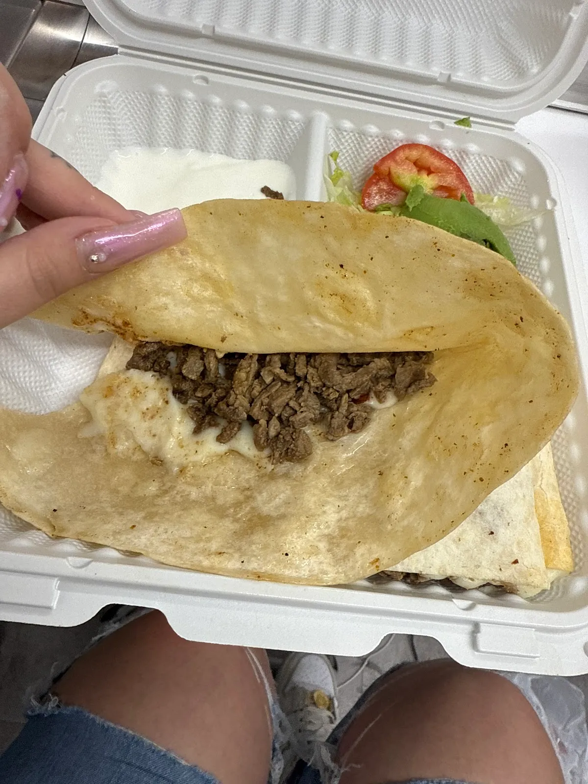 A grilled quesadilla with asada and melted cheese in a takeout container with avocado and tomato